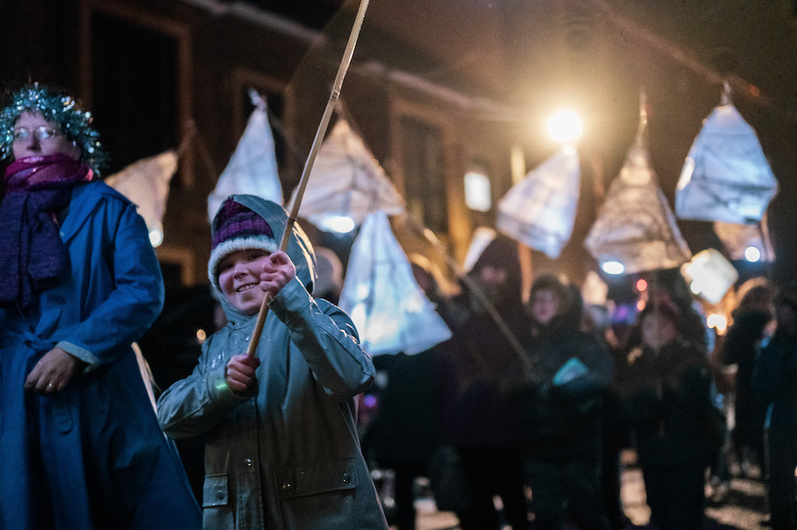 several people of all ages wrapped up in winter clothing parading outdoors with hand held paper lanterns.